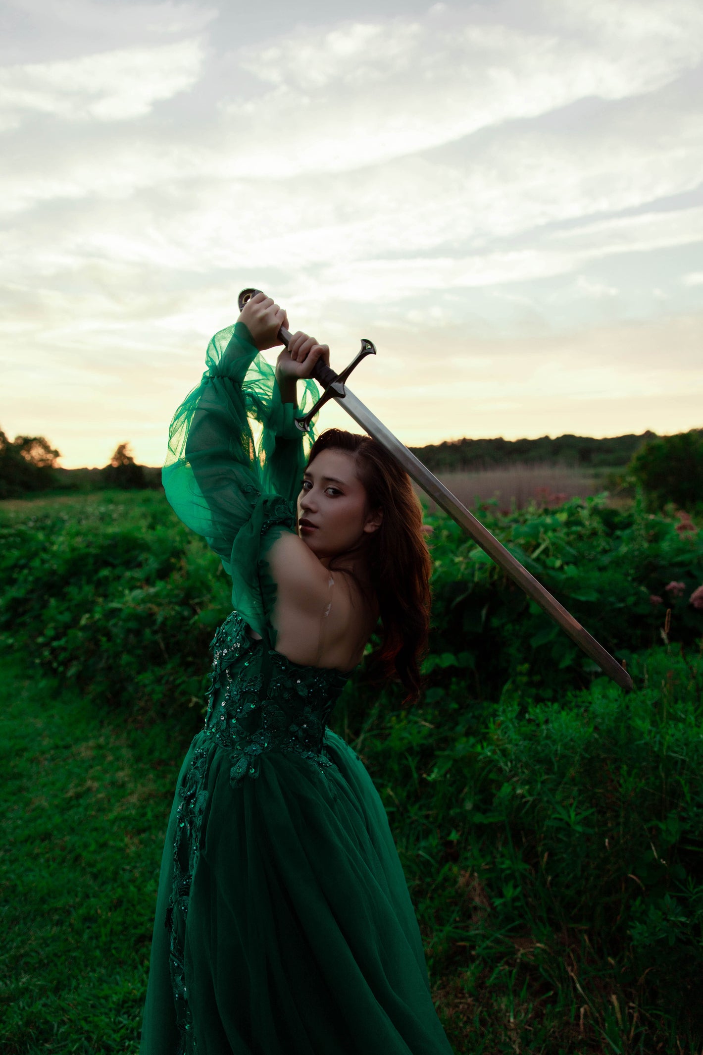 Woman in a green dress holding a sword in a grassy field with a cloudy sky.