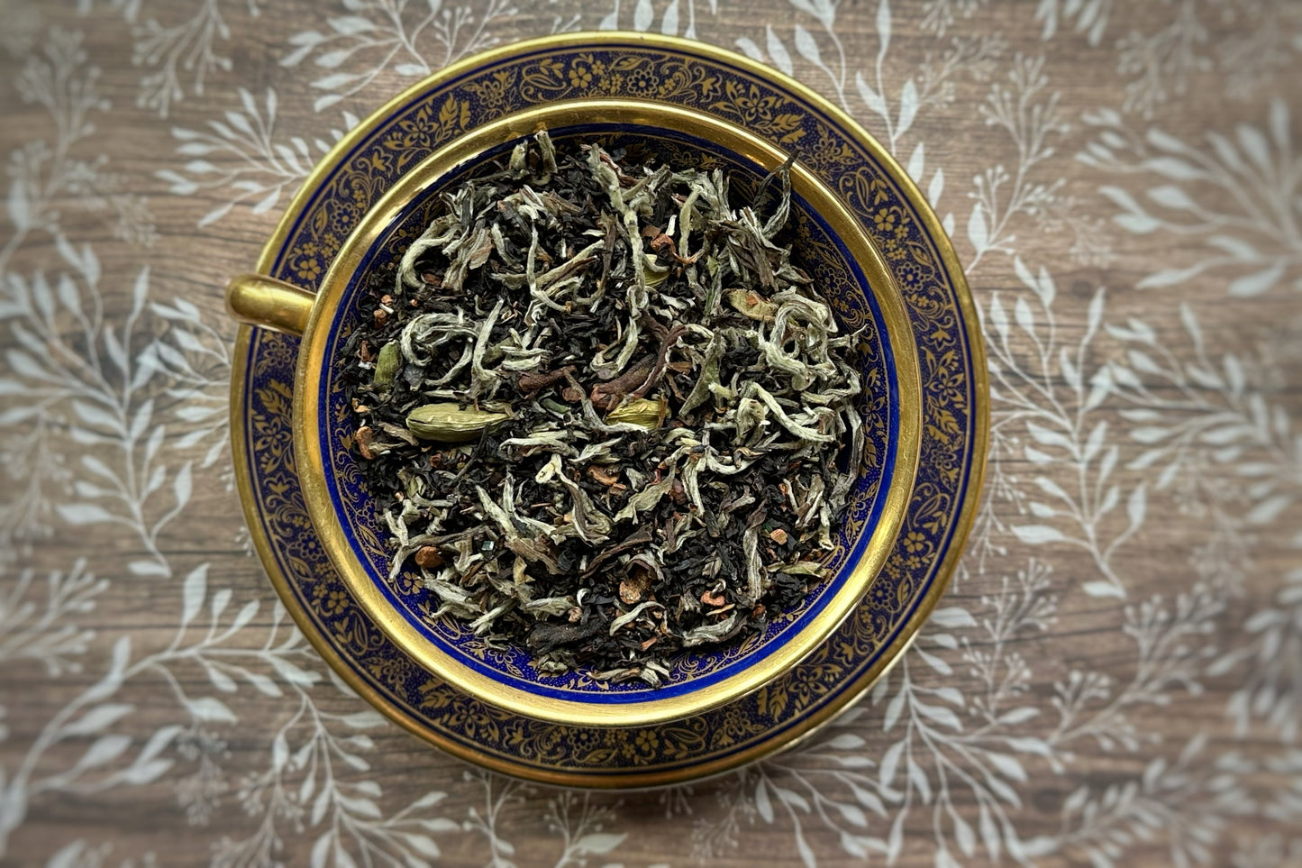 Dry tea leaves in a decorative blue and gold bowl on a wooden surface with leaf pattern