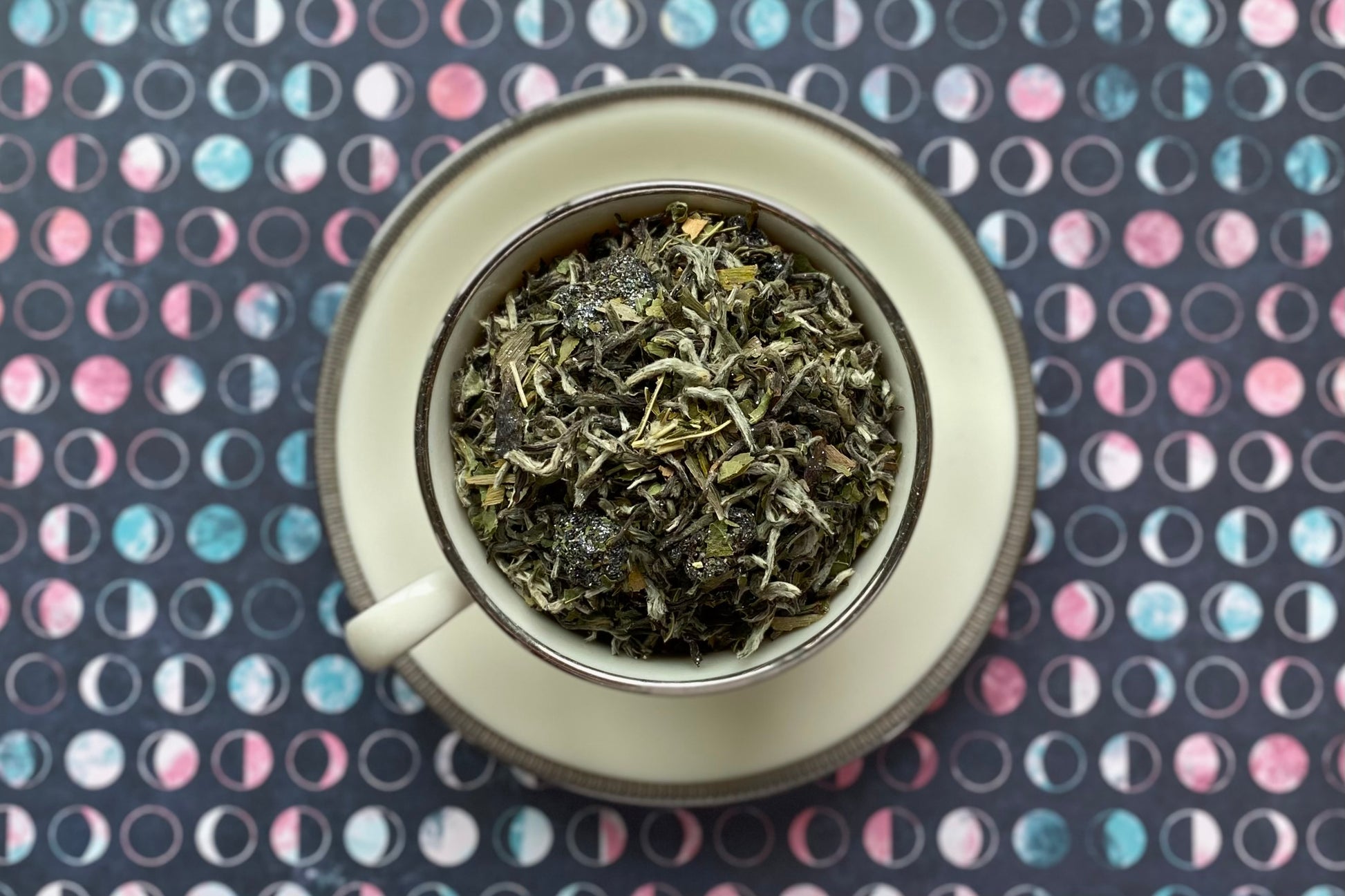 White tea with leaf and berry in white teacup with silver edges on a blue, white and pink moon phase background