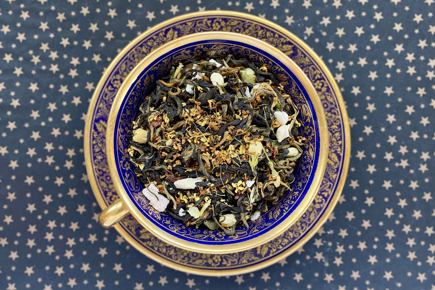 Blue and gold floral teacup full of gold-tipped tea leaves, flowers and peach pieces on starry backdrop
