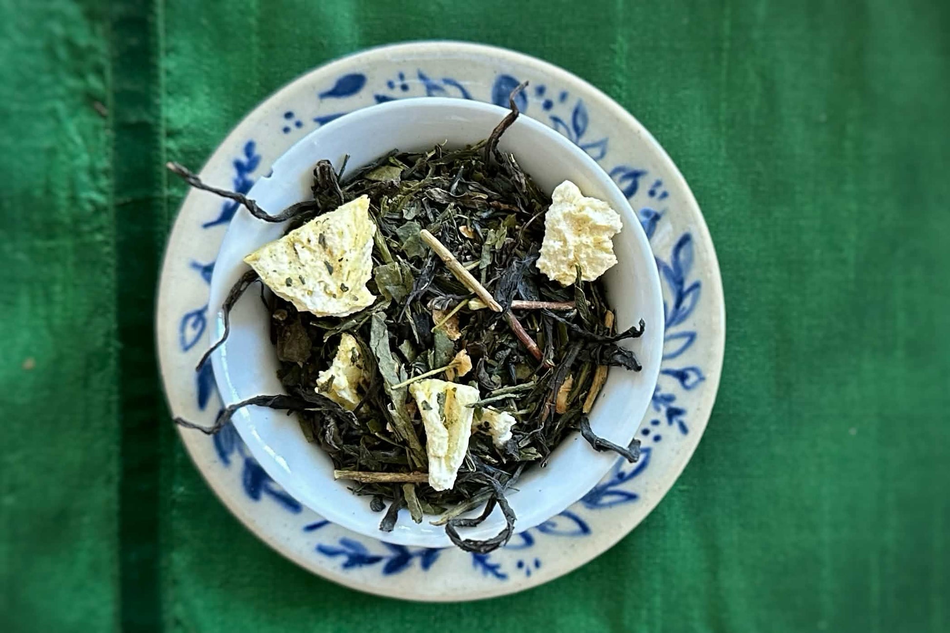 White teacup full of green tea yuzu peel and pineapple with wiry threads of fireweed creeping over the rim, all on a blue floral dish