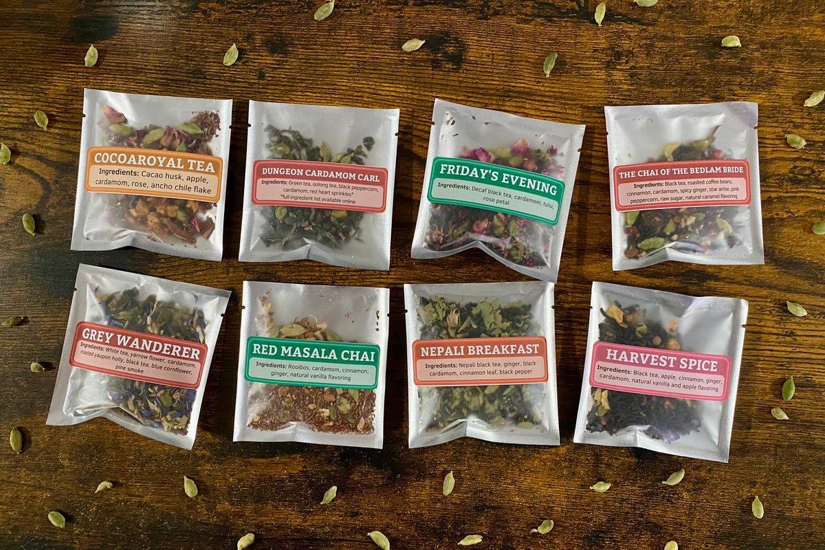 Eight individual tea bags arranged in two rows on a rustic wooden surface. Each clear resealable pouch has a colorful label naming a different tea variety: Cocoa Royal Tea, Dungeon Cardamom Carl, Friday's Evening, The Chai of the Bedlam Bride, Grey Wanderer, Red Masala Chai, Nepali Breakfast, and Harvest Spice.
