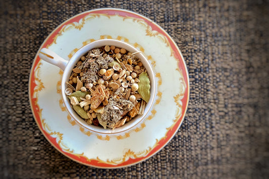 A cup of tea with a variety of spices and herbs on a saucer, including coriander, cardamom, ginger, and lemon peel.