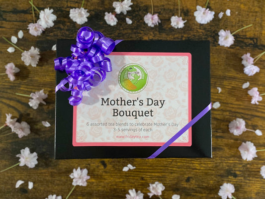 Gift box with 'Mother's Day Bouquet' label and purple bow on a wooden surface with scattered flowers.