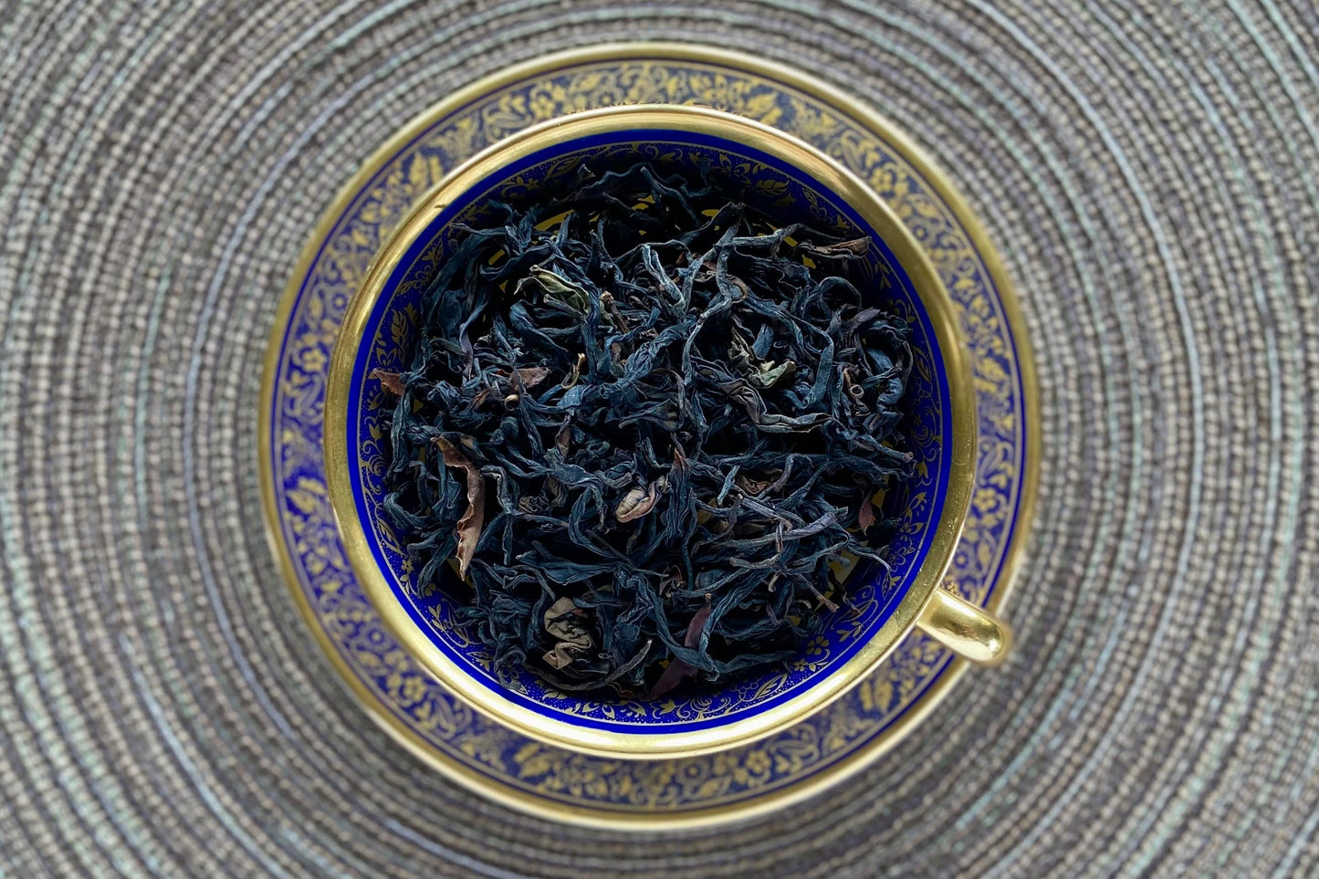 Blue and gold teacup full of wiry black tea with purple tips