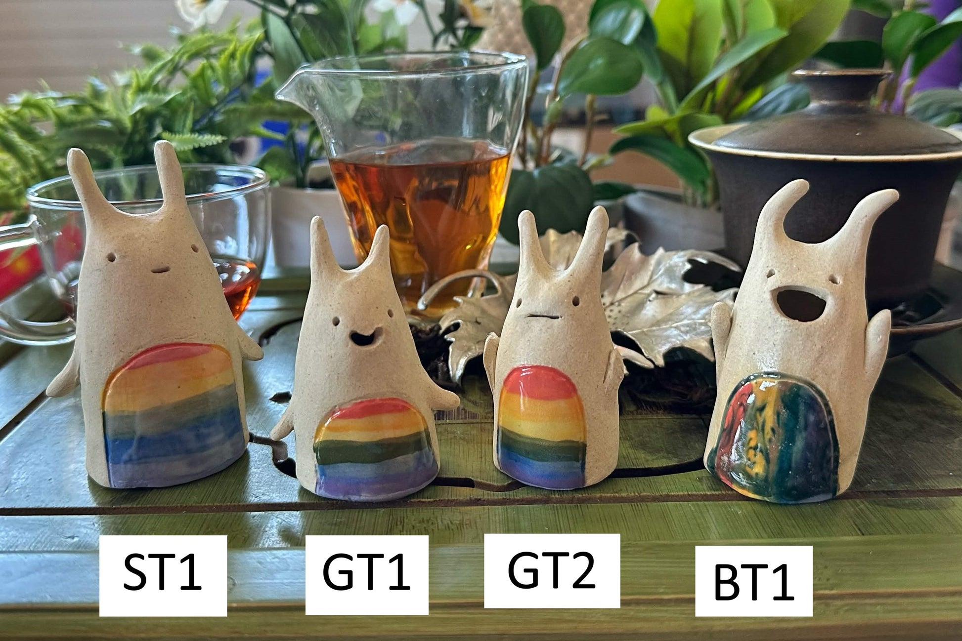 Four grinning horned blob tea pets with rainbow stripes on their bodies numbered ST1, GT1, GT2, BT1