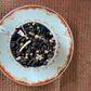 A cup of loose tea leaves and spices, including black peppercorns and coriander, is displayed on a saucer with a patterned edge. The tea setup is placed on a textured surface.