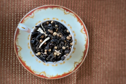 A cup of loose tea leaves and spices, including black peppercorns and coriander, is displayed on a saucer with a patterned edge. The tea setup is placed on a textured surface.